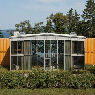 Modern house with large glass windows and orange exterior walls, surrounded by trees and a clear sky.