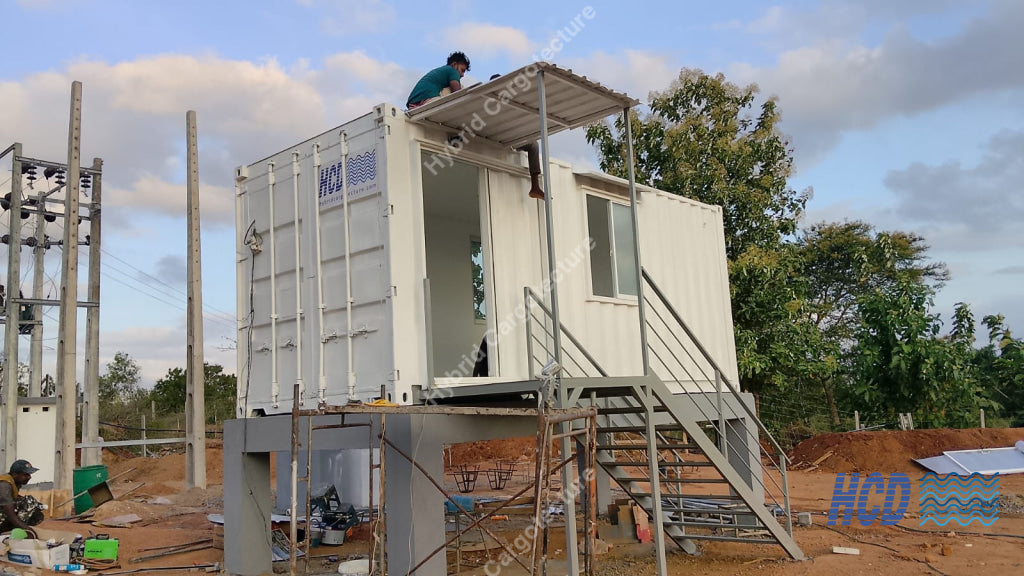 A white shipping container elevated on a metal frame with an attached staircase, set outdoors in a construction or utility site under a cloudy sky. A person is visible standing on the container roof.