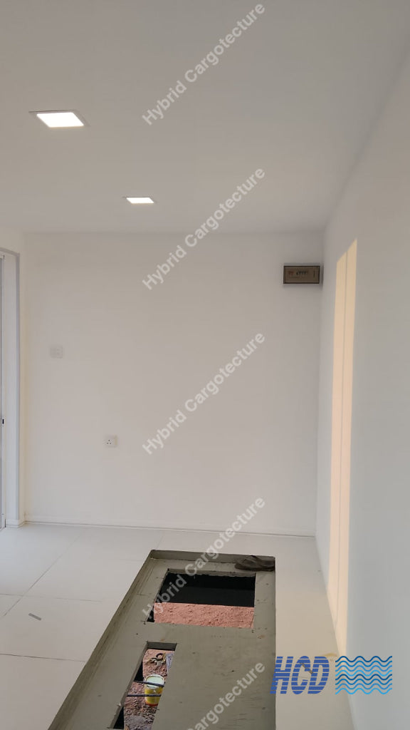 An empty white room with a rectangular floor opening showing construction materials inside, under recessed ceiling lights in a shipping container office.