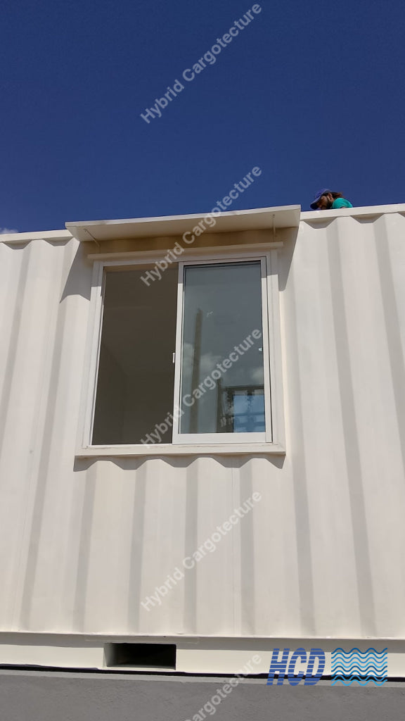 A white corrugated modular container home with a large window under a blue sky, with a person visible on the roof.