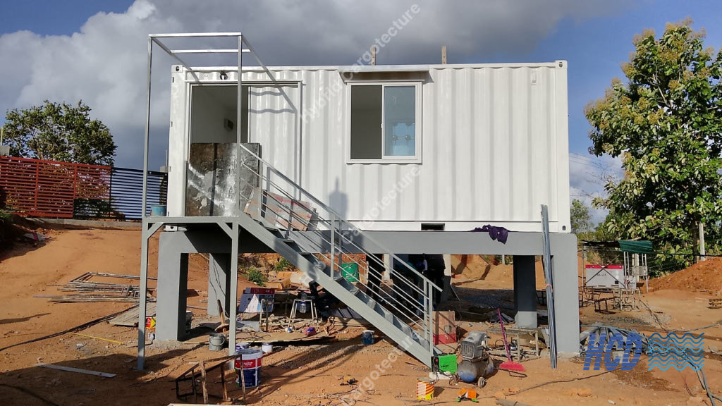 A white shipping container elevated on a metal support frame with an attached staircase, set outdoors on a dirt lot with trees, sky, and construction materials visible around it.