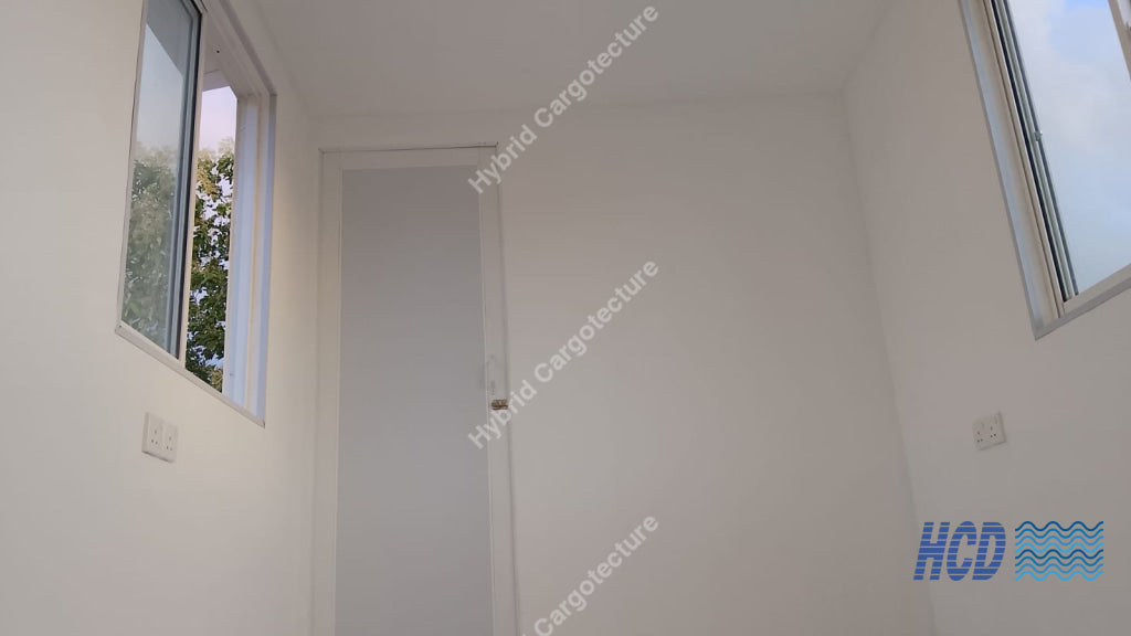 An empty white room with a closed white door in the center, flanked by two tall windows showing green trees and blue sky outside in a shipping container office.