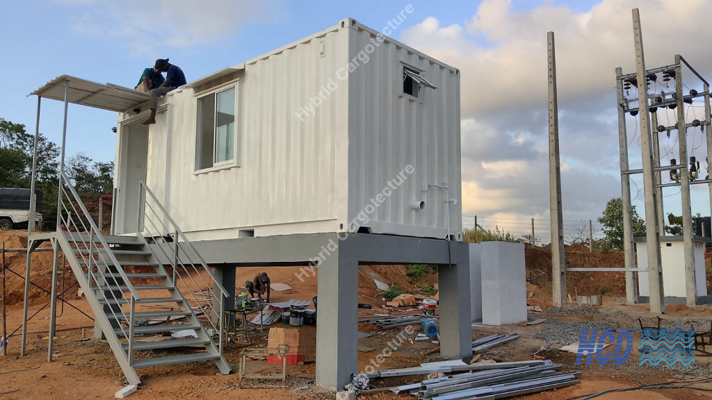 A white shipping container living room elevated on a metal frame, set outdoors on a construction site with a staircase, cloudy sky, and utility poles in the background.