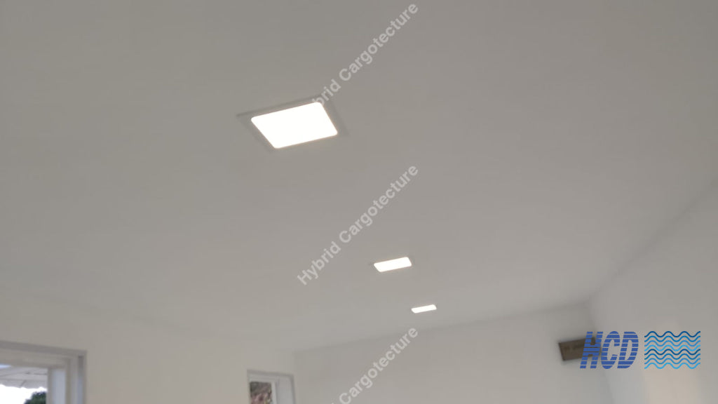 A white ceiling with three recessed square LED light fixtures installed at different heights in a shipping container office.