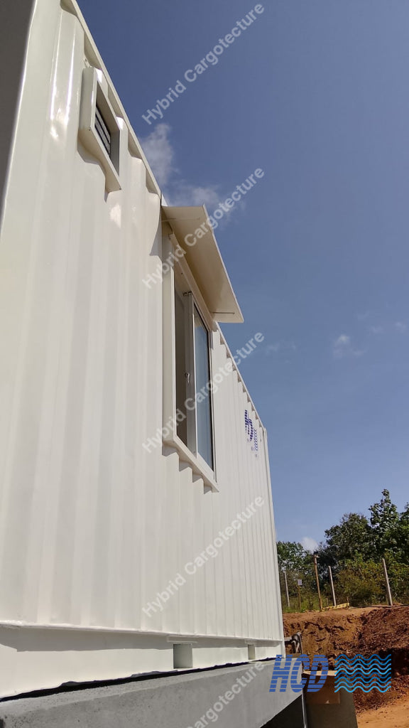 A white modular shipping container home with a corrugated metal exterior, featuring a window and a small vent, shown outdoors against a blue sky with some trees and dirt visible in the background.