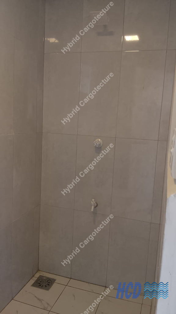 A tiled shower corner with light beige wall tiles, a floor drain, and a small white fixture on the wall in a shipping container office.