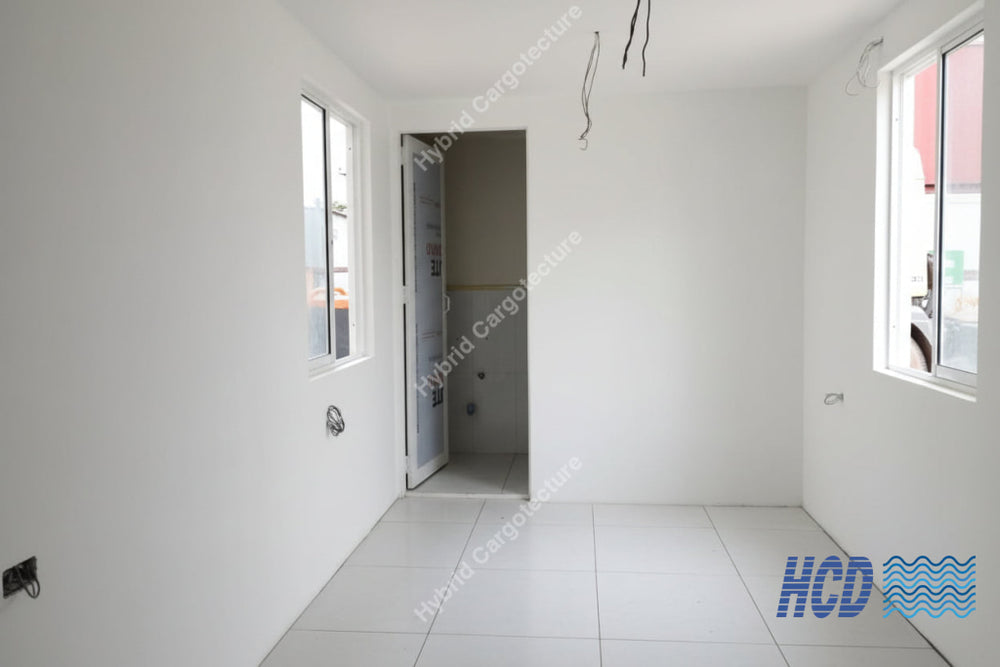 An unfinished room with white walls and floor, showing construction debris, an open doorway, exposed ceiling wires, windows, and a red and black shop vacuum cleaner on the floor.
