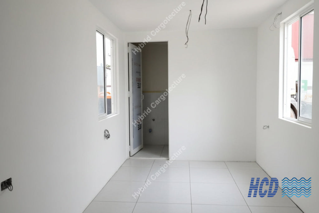 An unfinished room with white walls and floor, showing construction debris, an open doorway, exposed ceiling wires, windows, and a red and black shop vacuum cleaner on the floor.