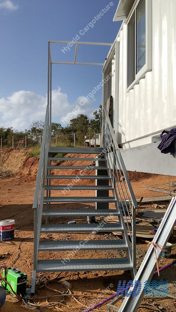 A galvanized steel outdoor staircase with handrails installed next to a white modular home on a dirt construction site under a blue sky.
