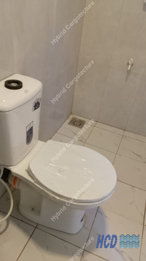 A white ceramic toilet with a closed lid, photographed in a tiled bathroom with light beige walls and floor in a shipping container office.