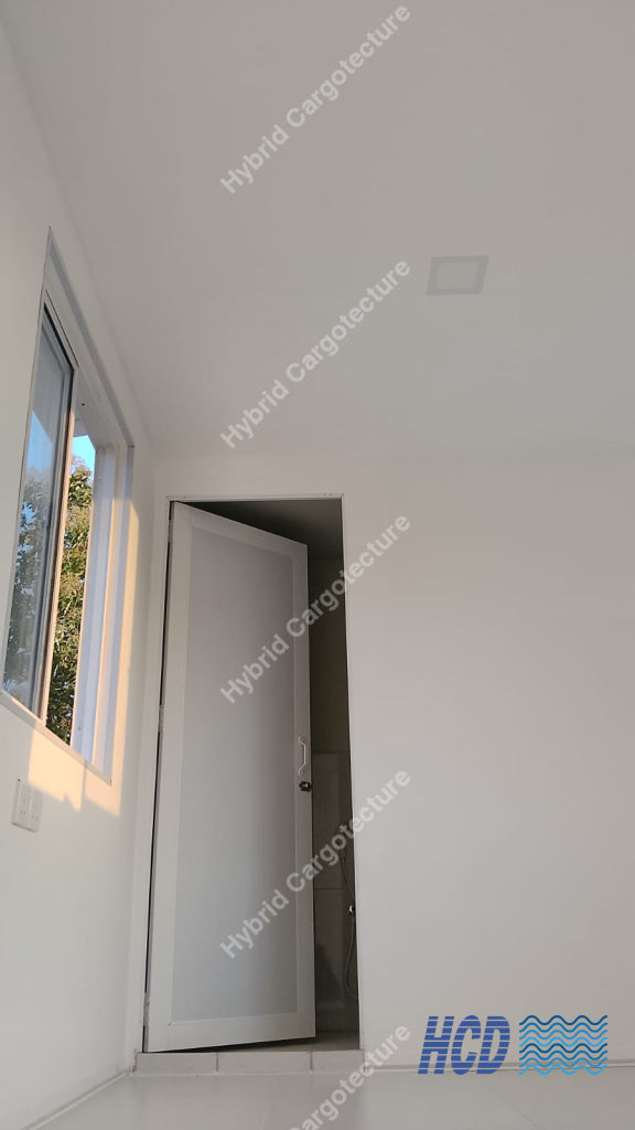 A white interior door with a dark handle, shown slightly open in a minimalist white room with a window on the left in a shipping container office.