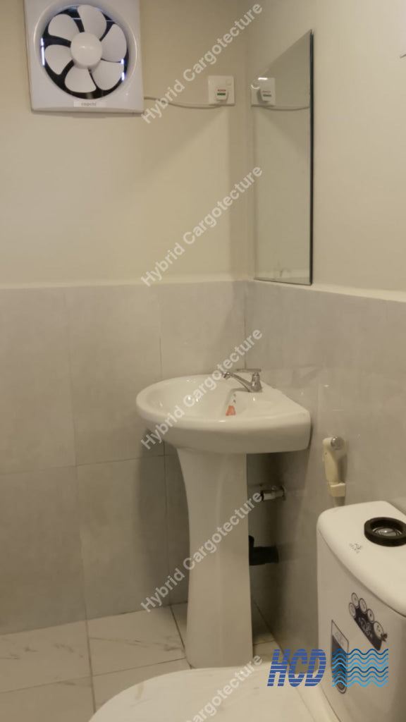 A compact bathroom corner with a white pedestal sink, toilet, wall mirror, tiled walls, and a wall-mounted exhaust fan in a shipping container office.