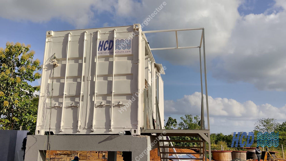 A large white shipping container-style unit with a metal frame structure on top, photographed outdoors under a cloudy blue sky. Trees and a dirt ground are visible around it, and a logo reading HCD is printed on the side.