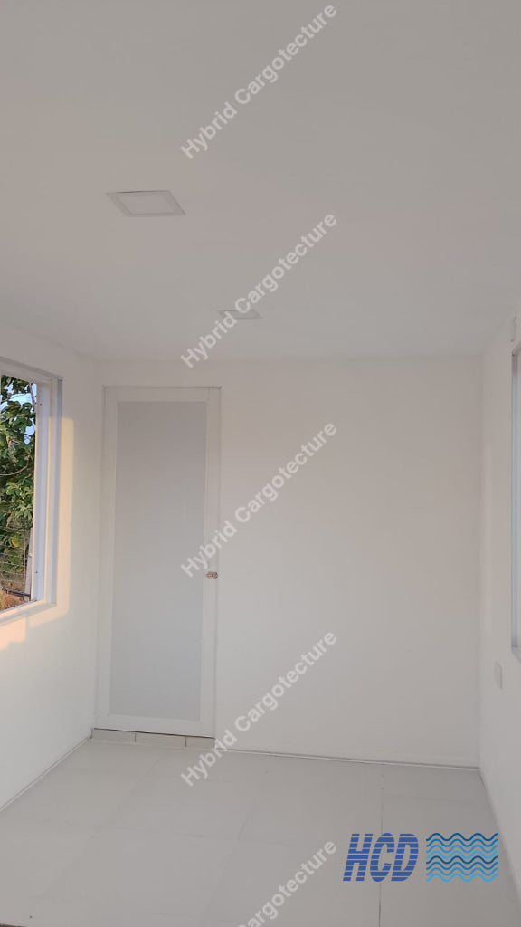 An empty, minimalist room with white walls, a white door, a window on the left showing greenery outside, and a light-colored tiled floor in a shipping container office.