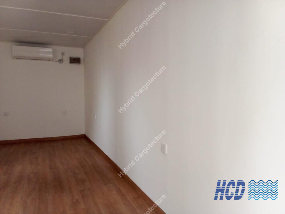 An empty room with white walls, a brown wood-look floor, and a white ceiling, featuring an air conditioner mounted on the left wall.