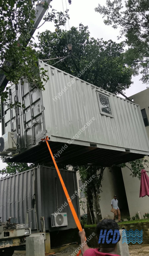 A large white shipping container being lifted by a crane outdoors, with an orange strap attached to its underside. Another container sits below on a truck bed, and a person in a pink shirt is visible in the foreground holding the strap.