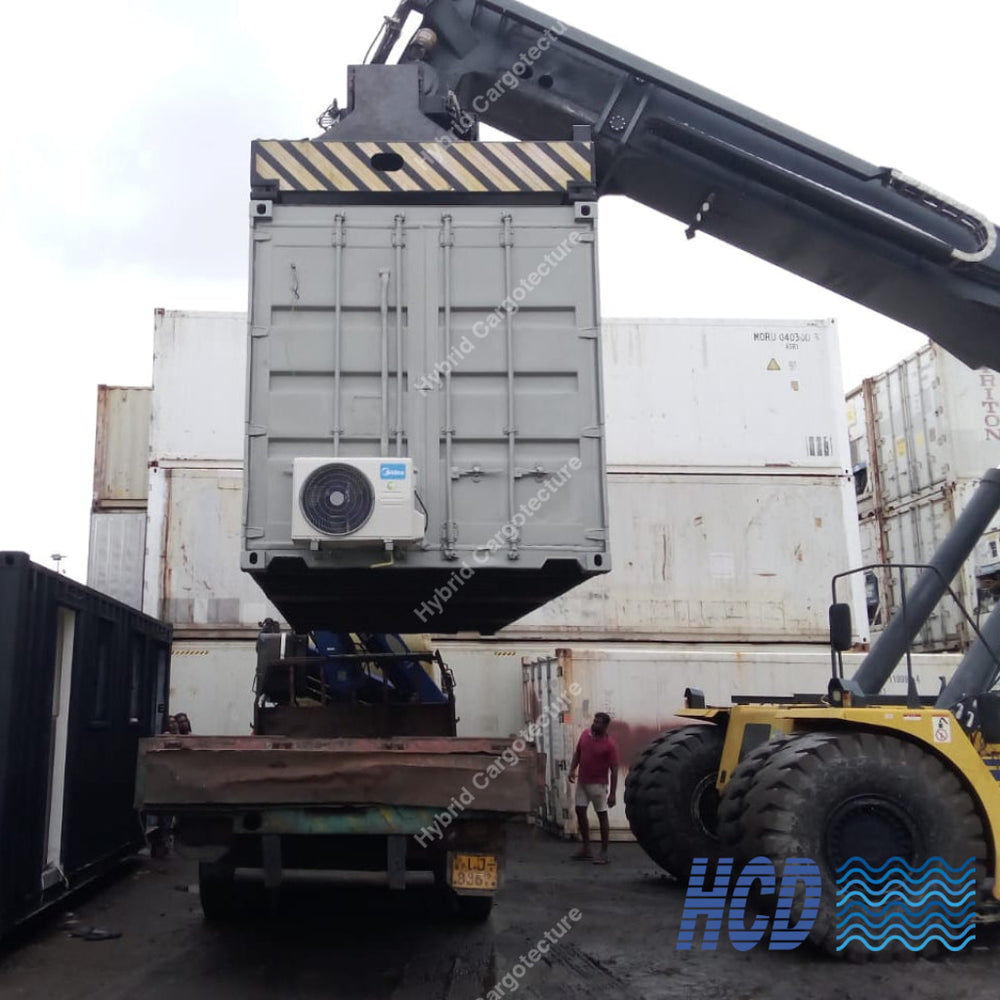 A large gray shipping container being lifted by a yellow crane attached to a truck in an outdoor industrial yard, with workers and other containers visible in the background.