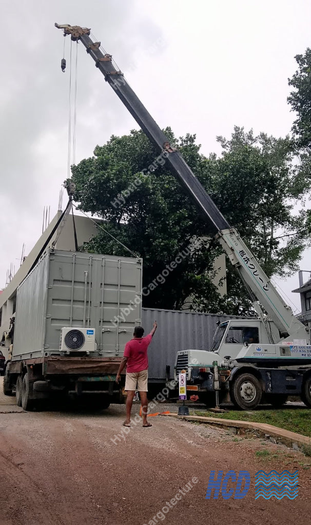 A large mobile crane truck parked beside a shipping container on an outdoor work site, with a person standing near the crane and container. Trees and an overcast sky are visible in the background.