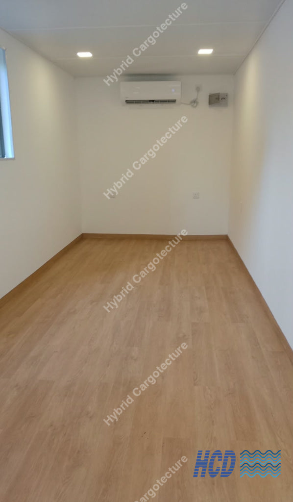 An empty room with light wood laminate flooring, white walls, a white ceiling with recessed lights, and a wall-mounted air conditioner. 