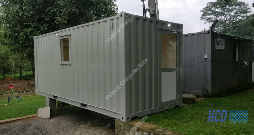 A light gray modular shipping container home elevated on a concrete base, situated outdoors on a grassy lawn next to another dark gray container unit.