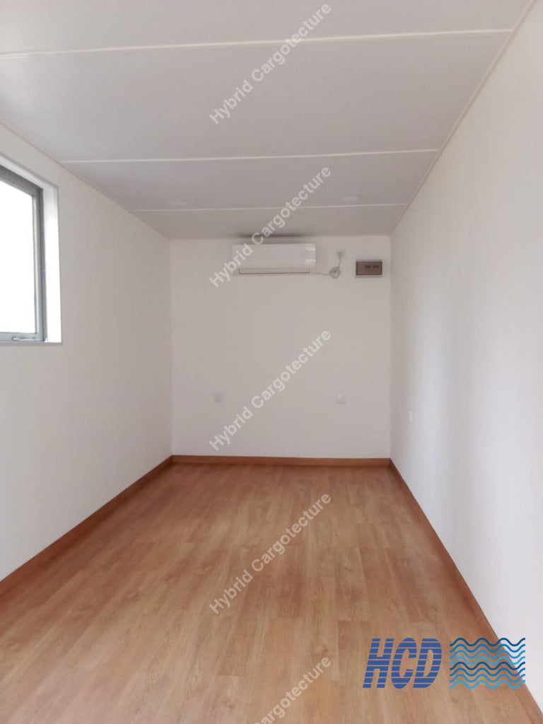 An empty room with light wood laminate flooring, white walls, a white ceiling, a window on the left, and a wall-mounted air conditioner.