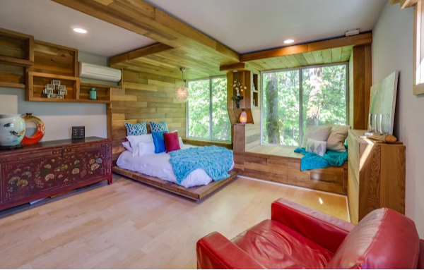 Wooden interior with a bed, red chair, and large windows.
