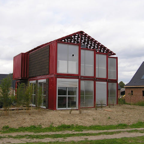 Modern house made from shipping containers with a red exterior and glass windows, set against a cloudy sky.