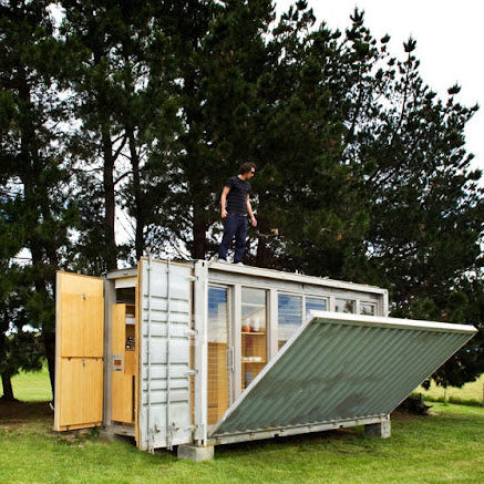 Small modular building made from shipping containers in a natural setting with trees and grass.