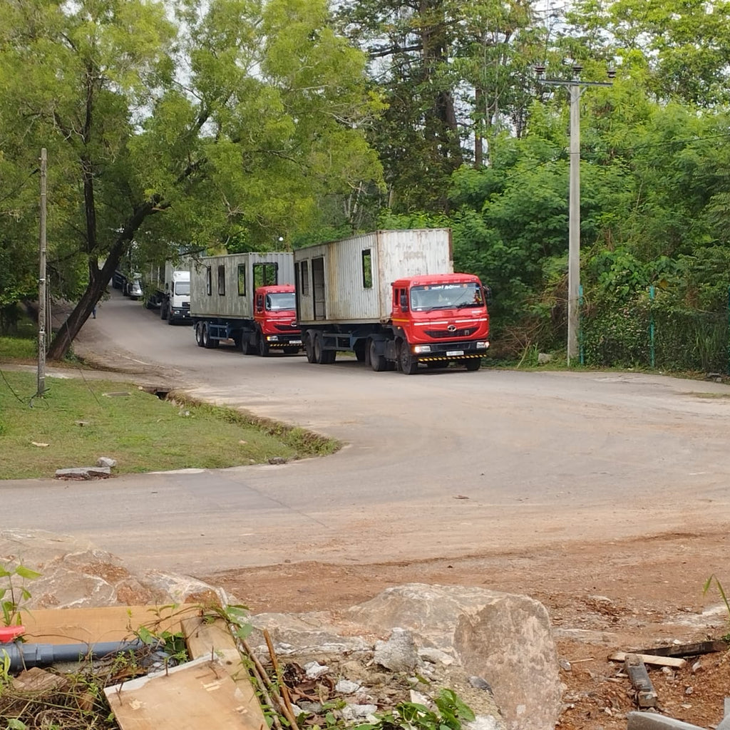 Red truck transporting a modular building on a rural road with trees and a fence in the background.
