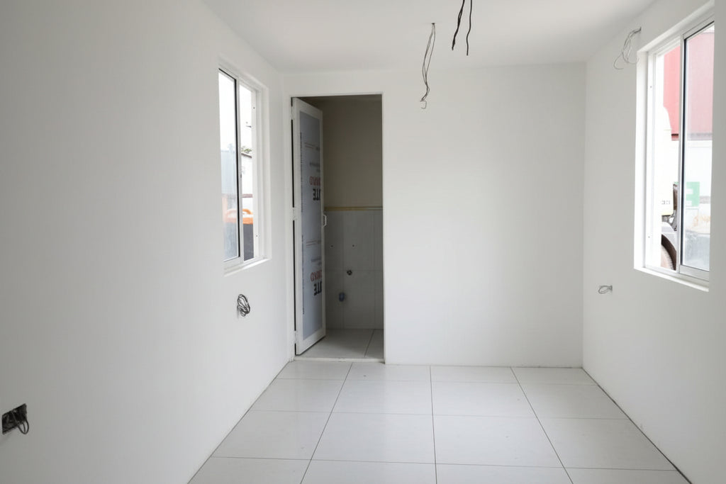 An unfinished room with white walls and floor, showing construction debris, an open doorway, exposed ceiling wires, windows, and a red and black shop vacuum cleaner on the floor.