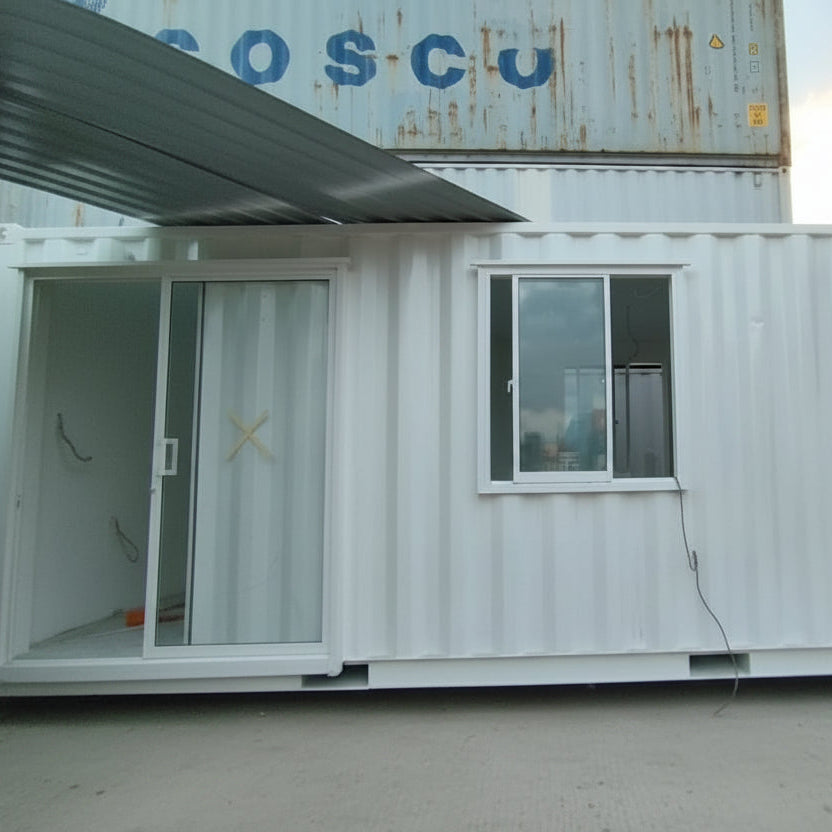 A white modular shipping container home with a sliding glass door and a window, set outdoors on a dirt ground with a black barrel in front.