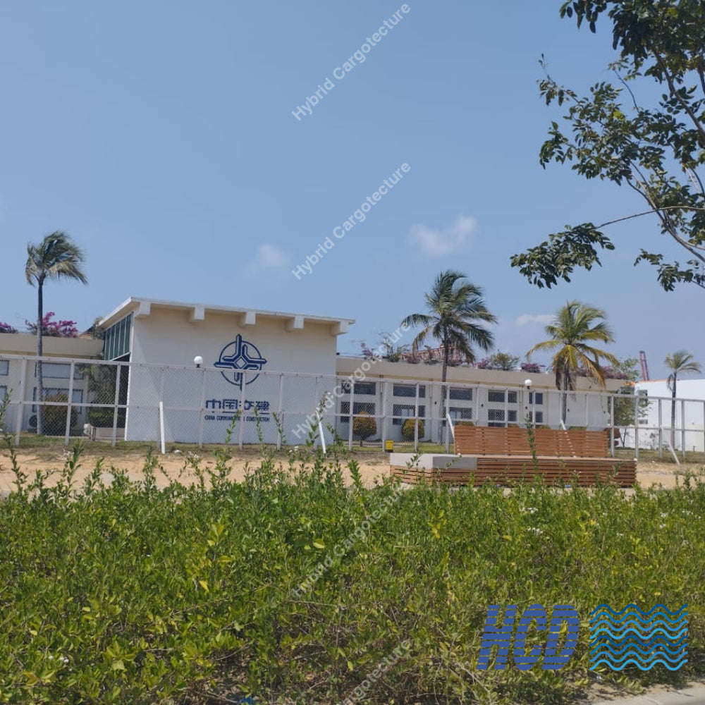 Building with a logo on the side, surrounded by palm trees and greenery under a clear blue sky.