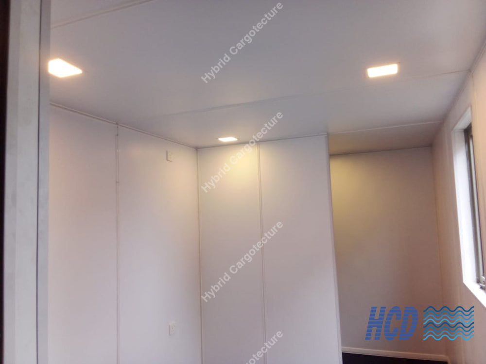 An interior room corner with white wall panels, a white ceiling, two recessed lights turned on, and a window on the right side.