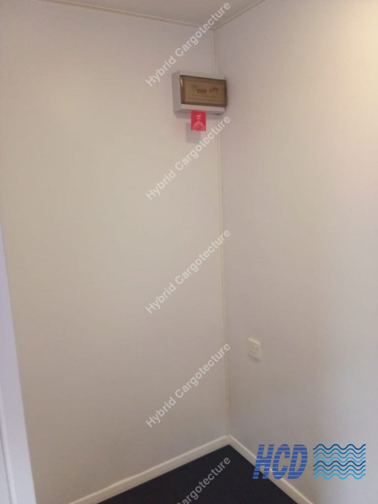 An empty white-walled room corner with a small wall-mounted box and red tag near the top, plus a dark floor covering.