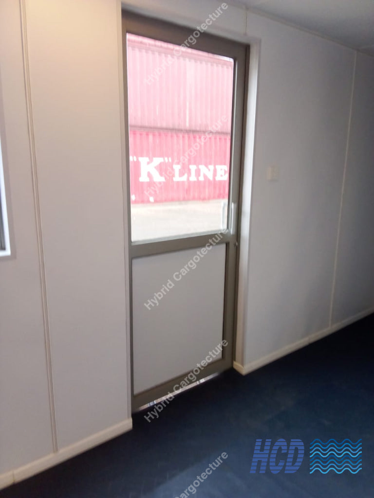 A modern modular office or warehouse door with a large window showing a red shipping container outside, set within light gray panel walls on a dark blue floor.