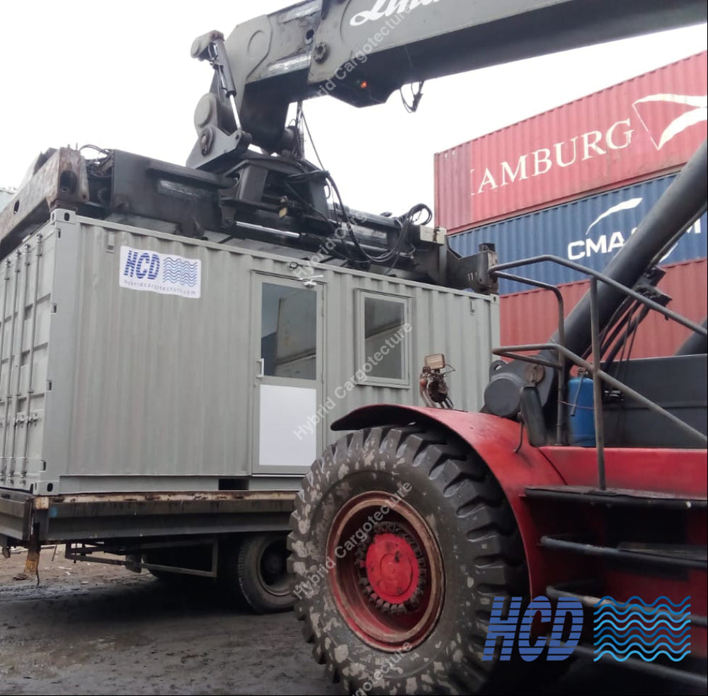 A large gray modular office container being lifted by a crane and pushed by a red tractor on a muddy outdoor industrial yard, with shipping containers and workers visible in the background.