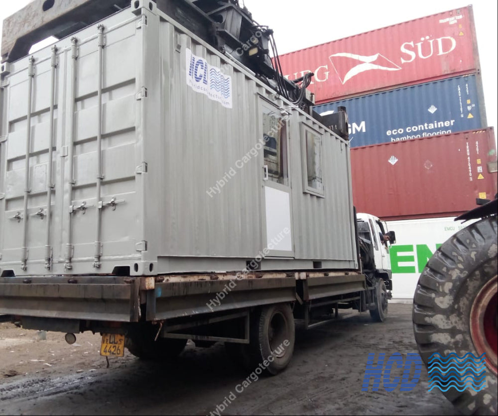 A large gray shipping container mounted on a flatbed truck, photographed in an outdoor industrial yard with stacked shipping containers and a tire visible in the foreground.