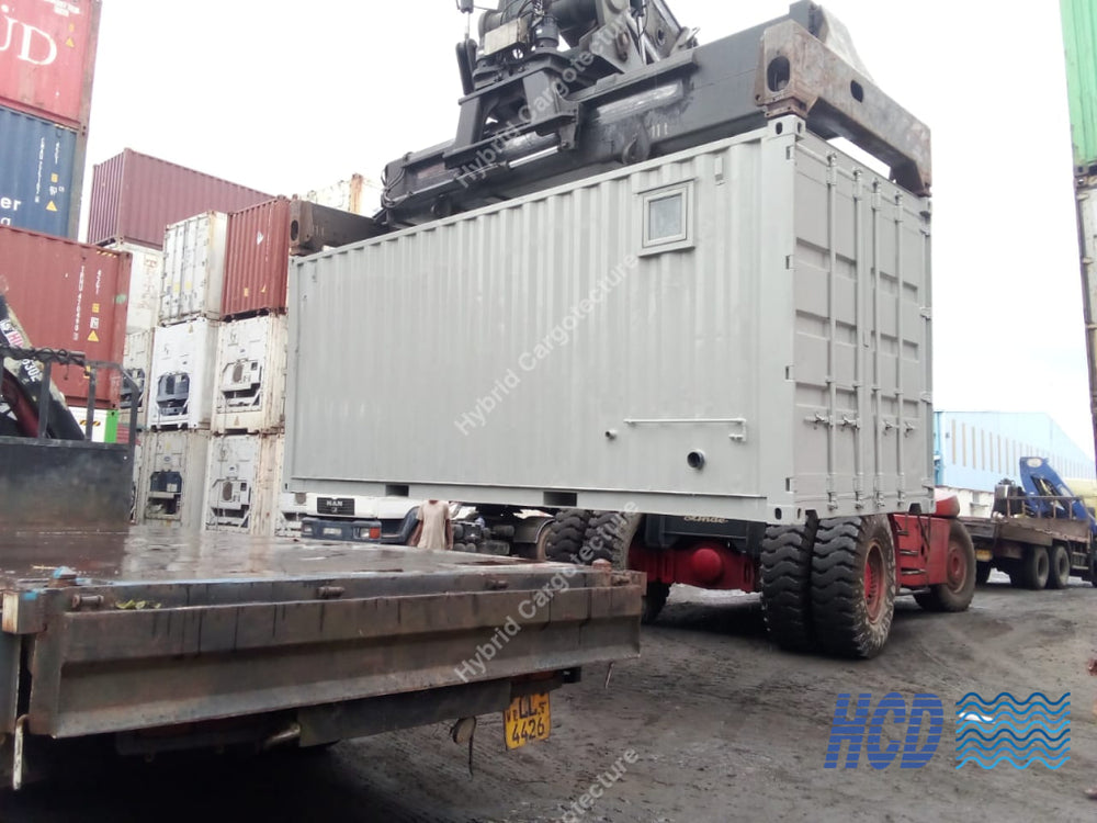 A large white shipping container mounted on a red flatbed trailer, being lifted by a crane in an outdoor port or warehouse setting with stacked containers visible in the background.