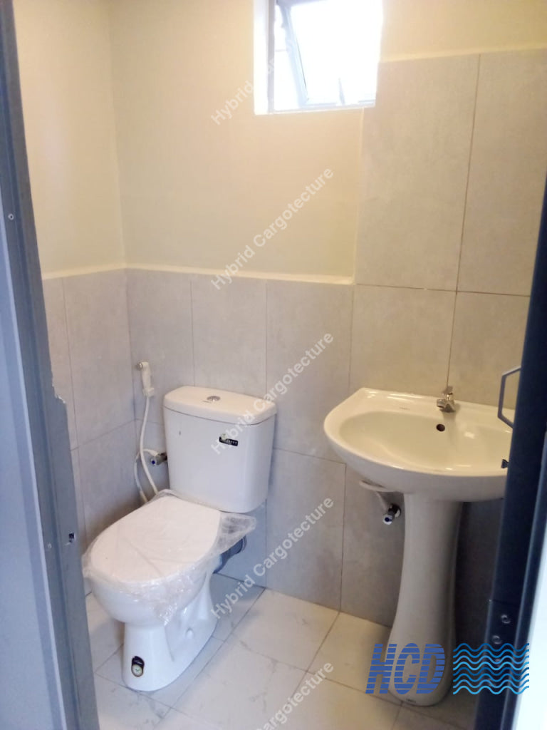 A compact bathroom corner with a white toilet wrapped in protective plastic, a white pedestal sink, light gray wall and floor tiles, and a small window near the ceiling.
