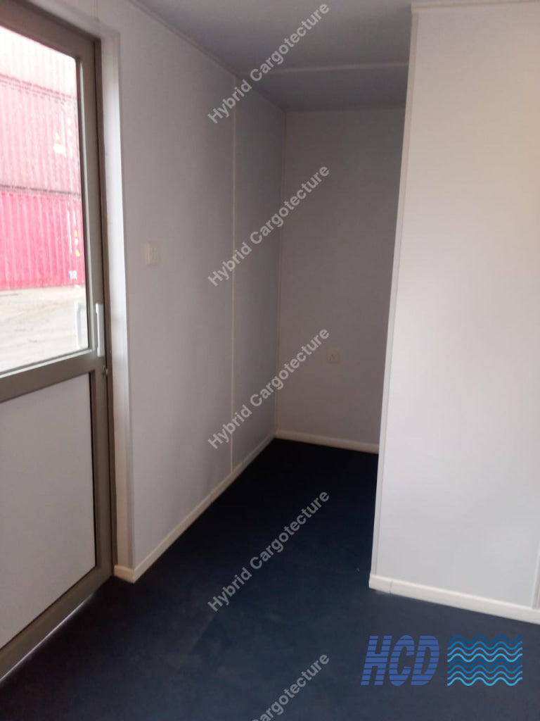 An empty room with white walls, a dark blue carpeted floor, and a glass door on the left side showing a red container outside.