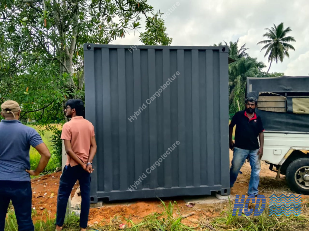 Three men standing around a large gray shipping container outdoors with trees and a truck in the background.